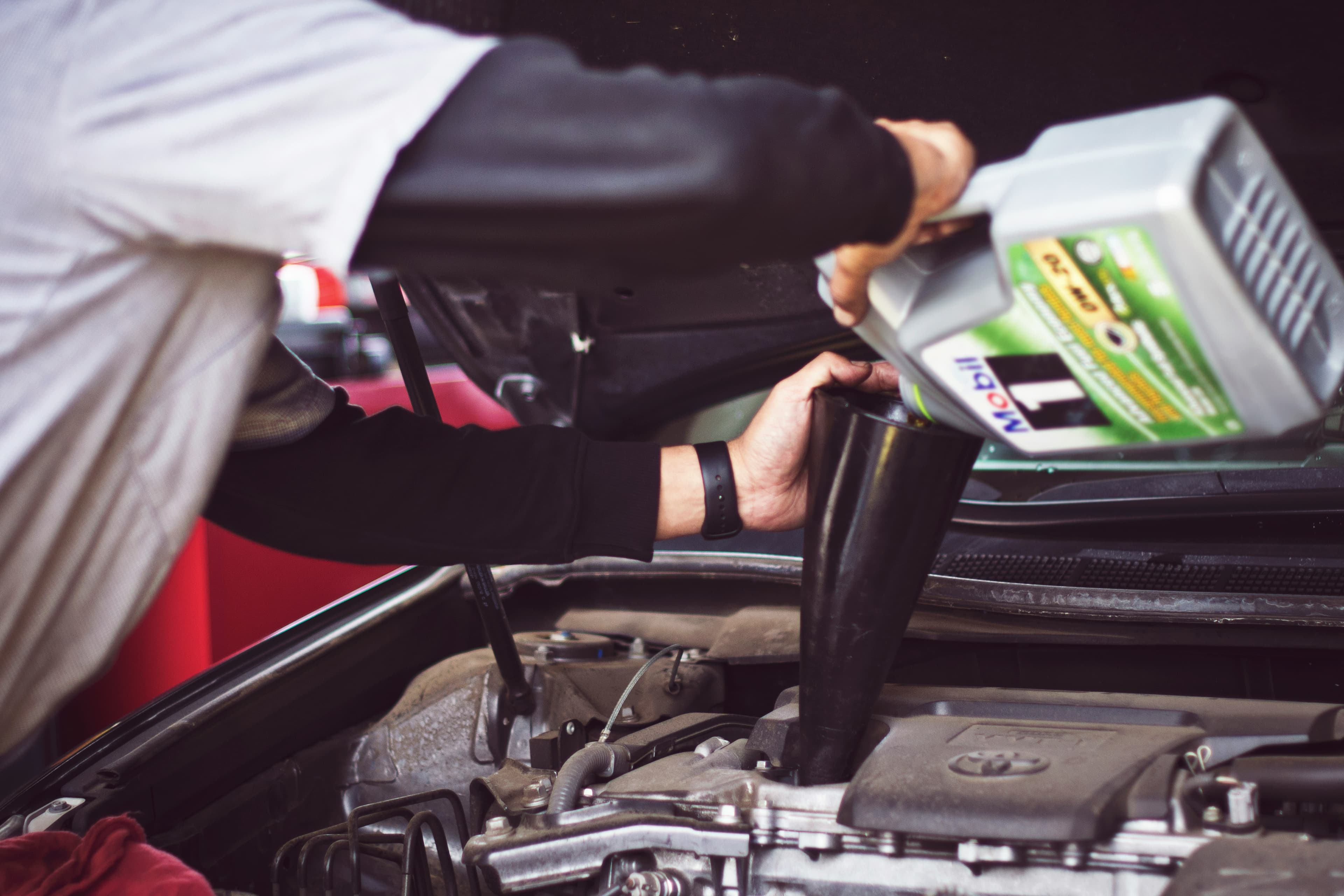 Picture of a mobile mechanic standing over a car with its hood open pouring oil into the car  